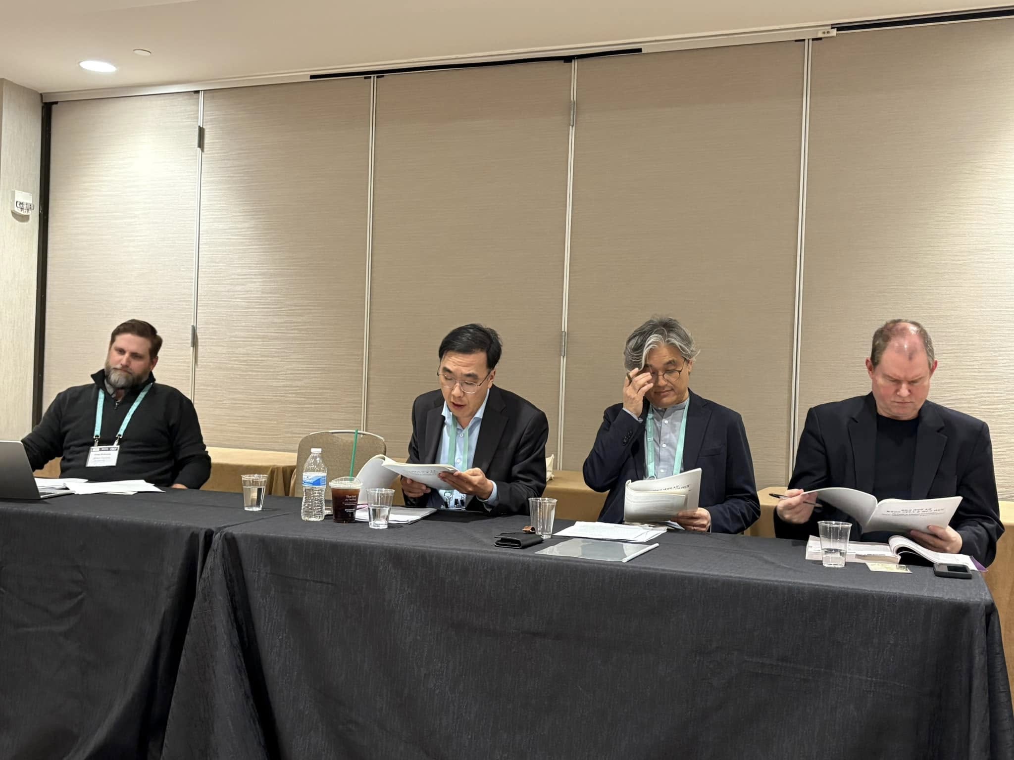 Four people seated side by side at a conference table with papers in front of them.